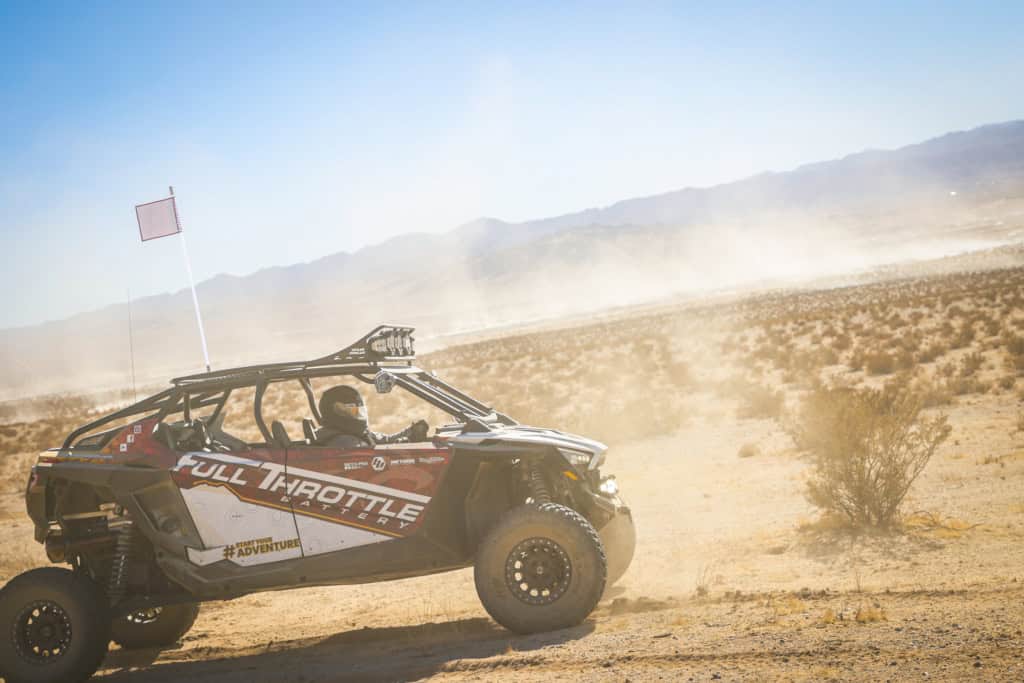 Off-road vehicle with "Full Throttle" branding speeds through a desert landscape, kicking up dust. A driver is visible inside, and a flag is attached to the vehicle.