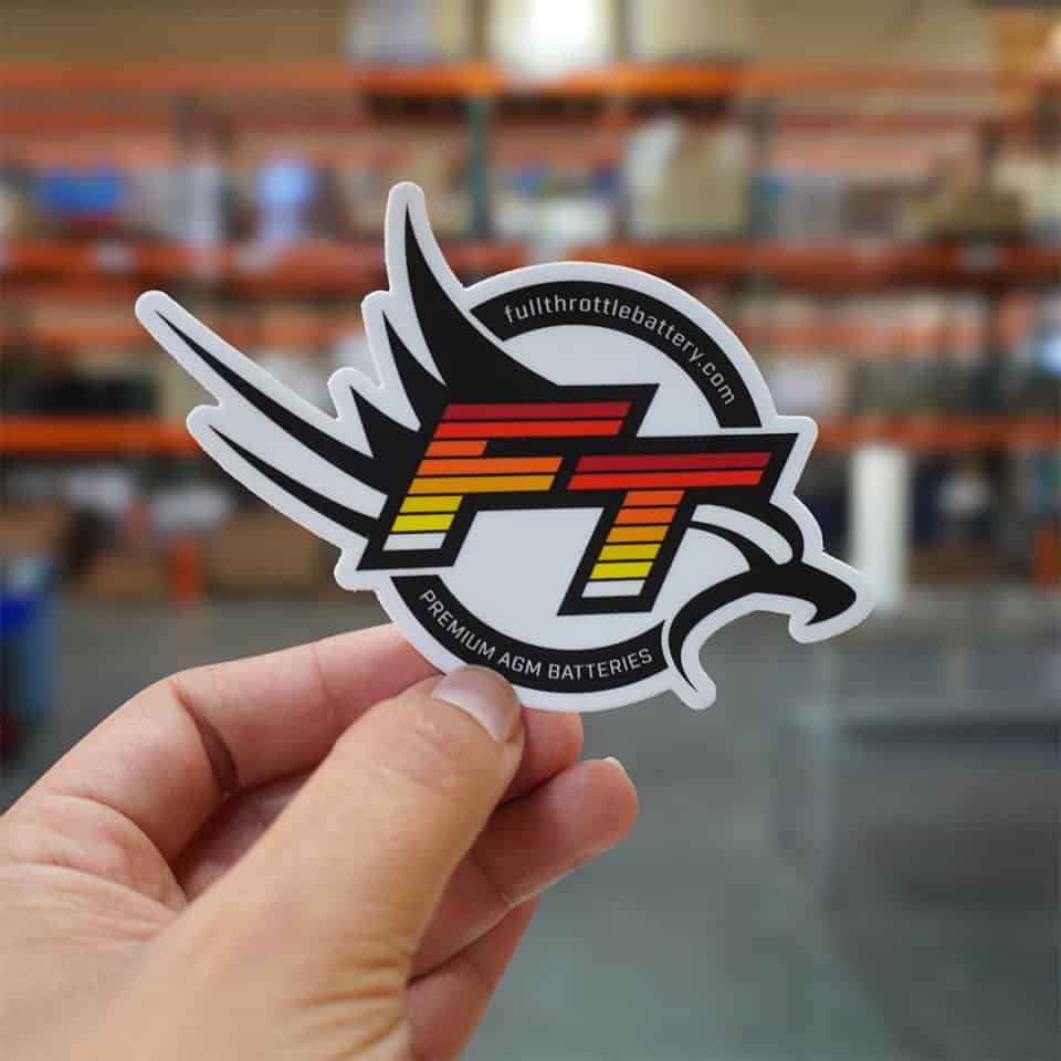 A hand holds an FT Sticker featuring the "FT" text and "fullthrottlebattery.com" along with a wing design, against a backdrop of blurred shelves.