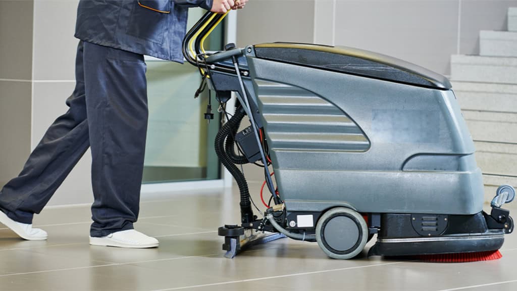 A person in work uniform operates a floor cleaning machine on a tile floor near a staircase.