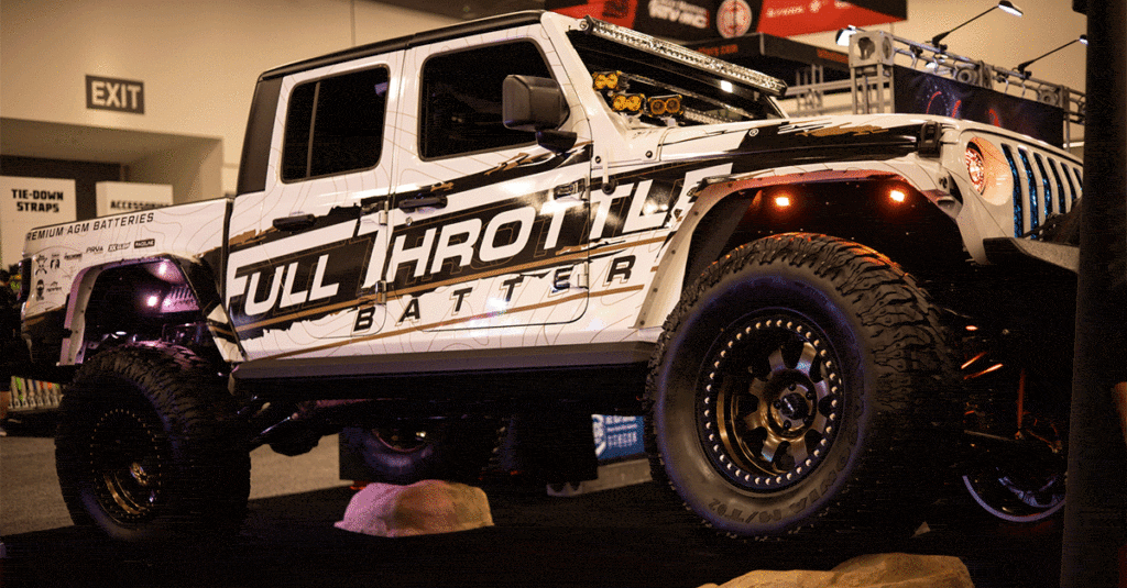 A modified white off-road vehicle with "full throttle" decals displayed at an indoor expo, equipped with large tires and additional lighting on top.