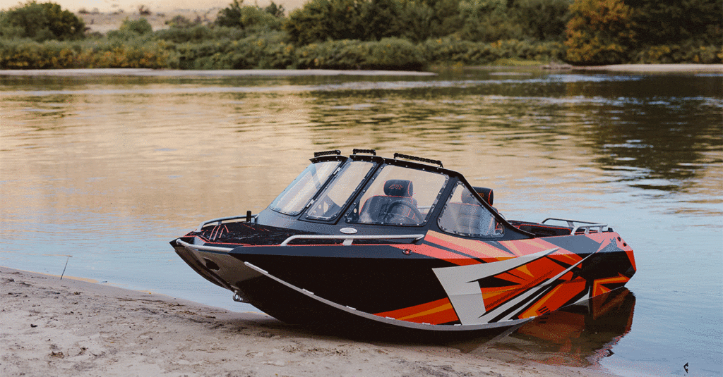 A black and orange speedboat parked on a sandy riverbank with calm waters and trees in the background.