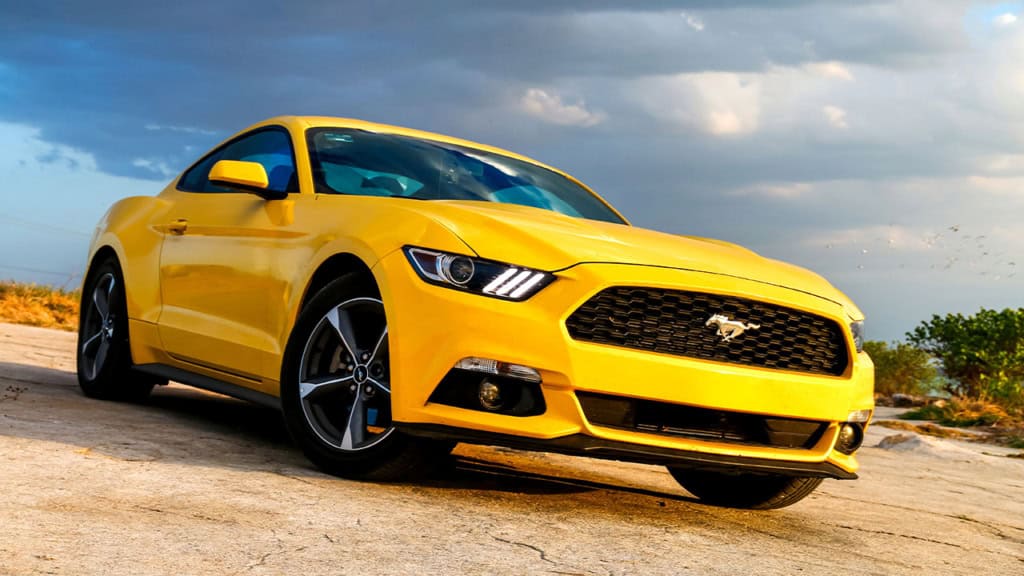 A yellow Ford Mustang coupe is parked outdoors on a concrete surface under a partly cloudy sky.