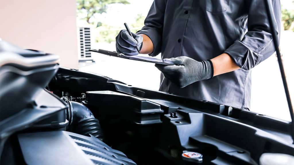 A mechanic wearing gloves inspects a car engine while making notes on a clipboard.