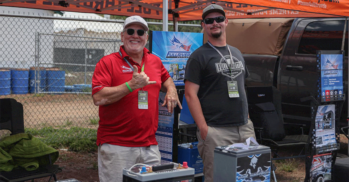 Two men standing at an outdoor event in a Full Throttle Battery booth, one giving a thumbs-up, surrounded by promotional banners and products on a table.