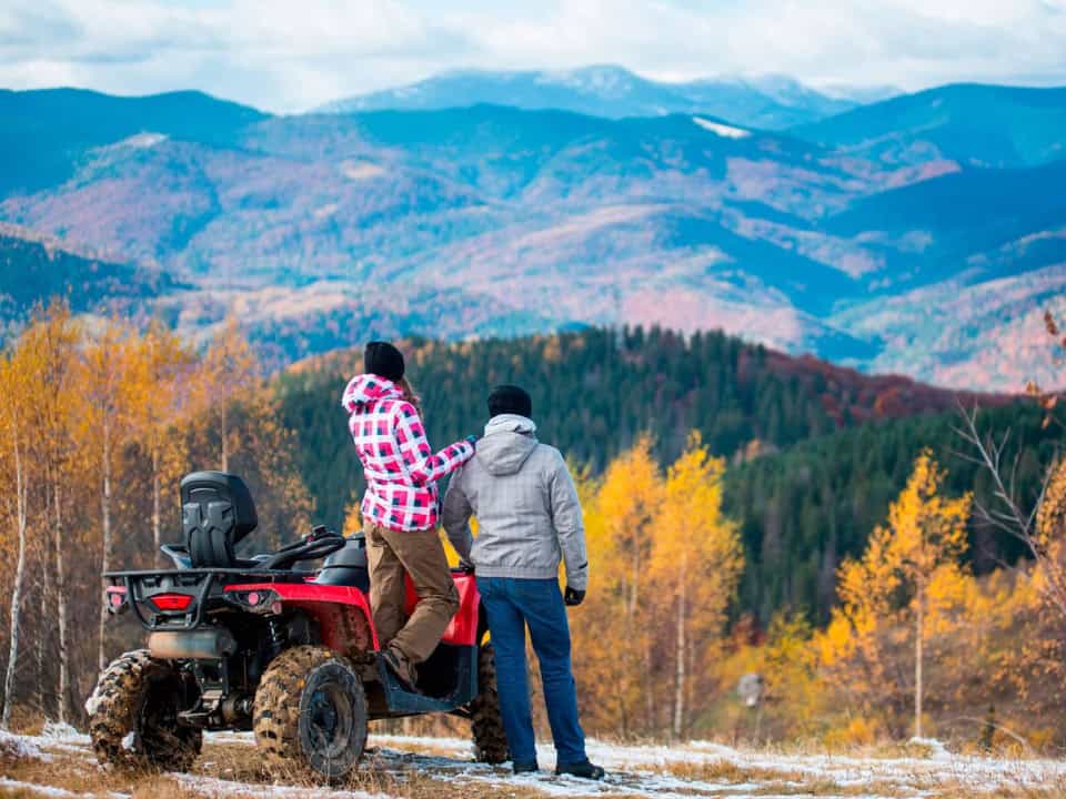 Two people in winter clothing stand by a red ATV, looking at a scenic view of mountains and autumn trees.