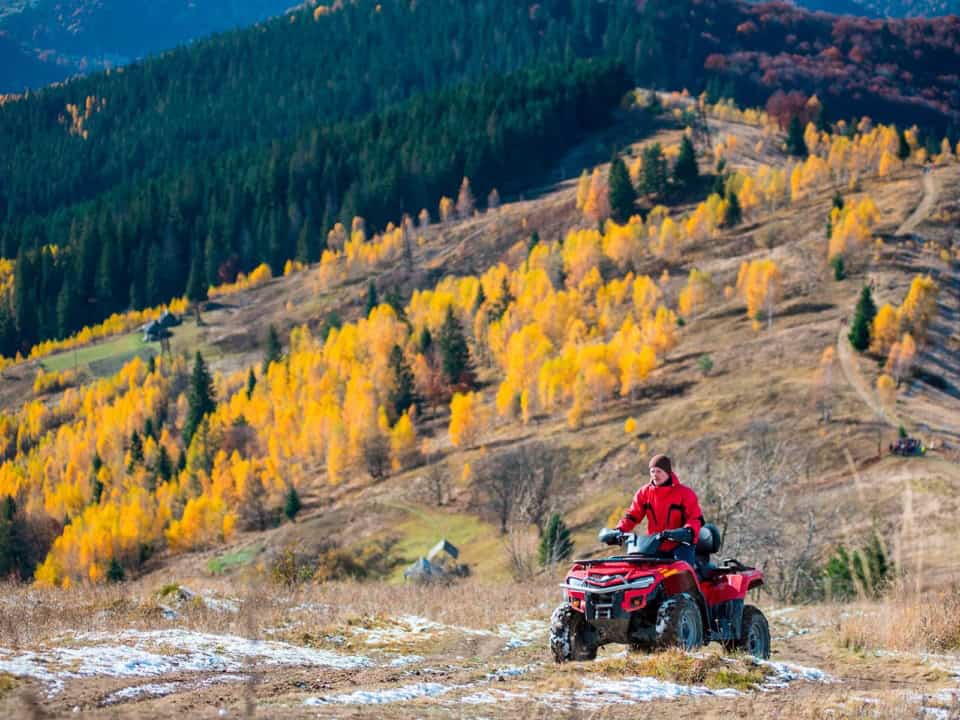 Person in a red jacket riding an ATV on a mountain path, surrounded by autumn foliage and distant hills.