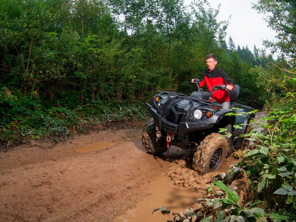 A person wearing a red and black jacket rides an ATV through a muddy path surrounded by dense green foliage.