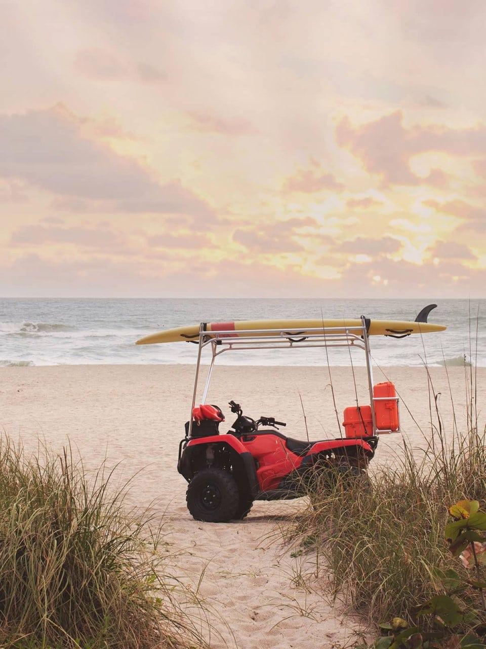 A red ATV with a surfboard on top is parked on a sandy beach path, surrounded by dune grass, with the ocean and a cloudy sky in the background.