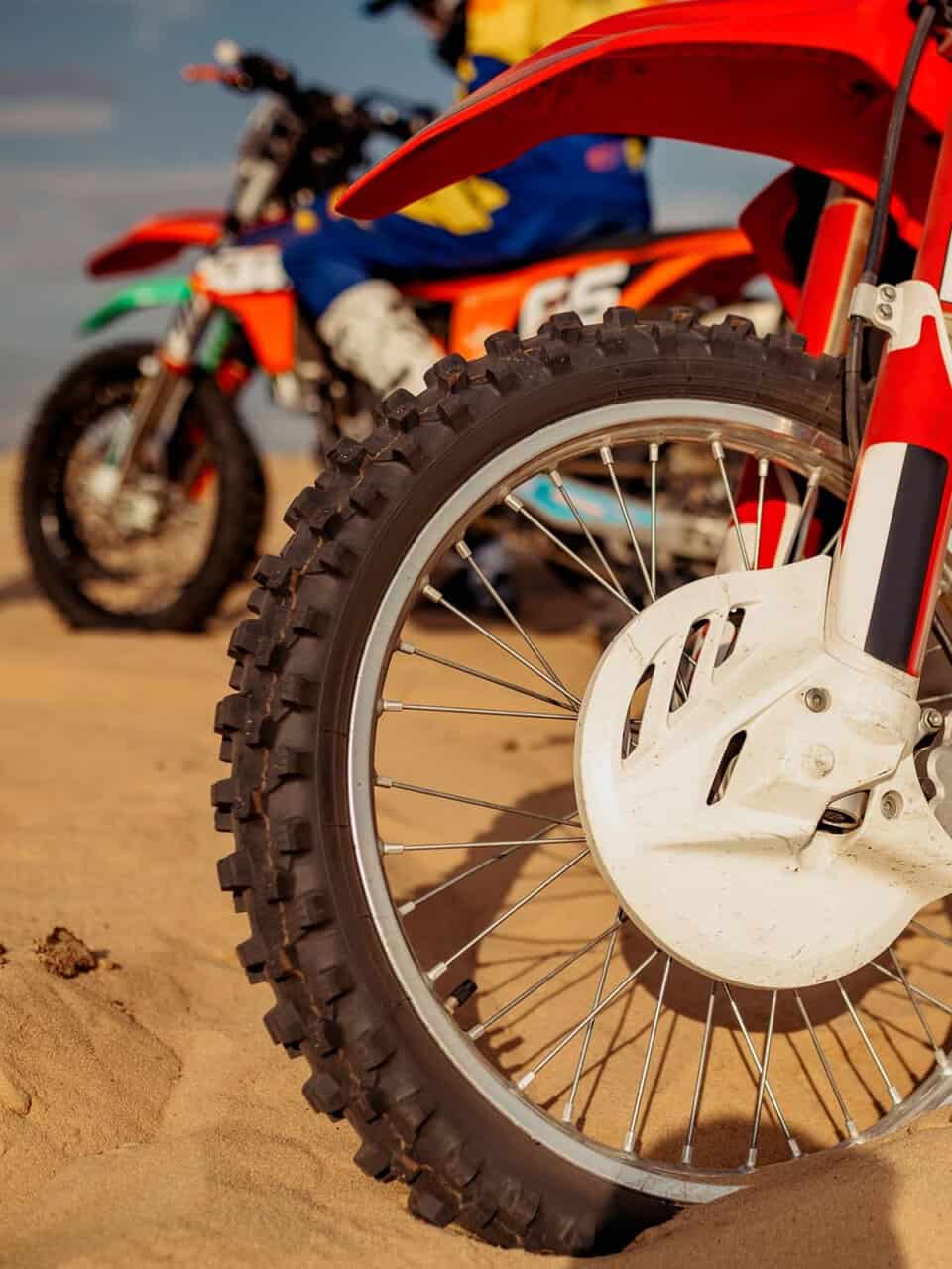 Close-up of a red dirt bike wheel on sandy terrain, with another dirt bike and rider in the background.