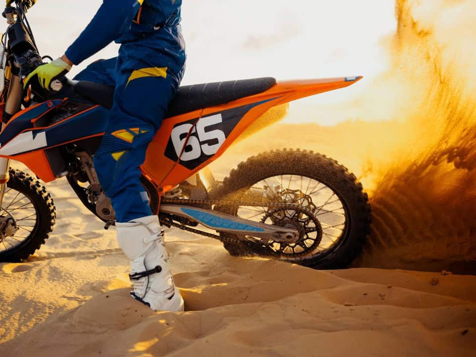 A motocross rider on a dirt bike with the number 65 kicks up sand while riding on a dune.