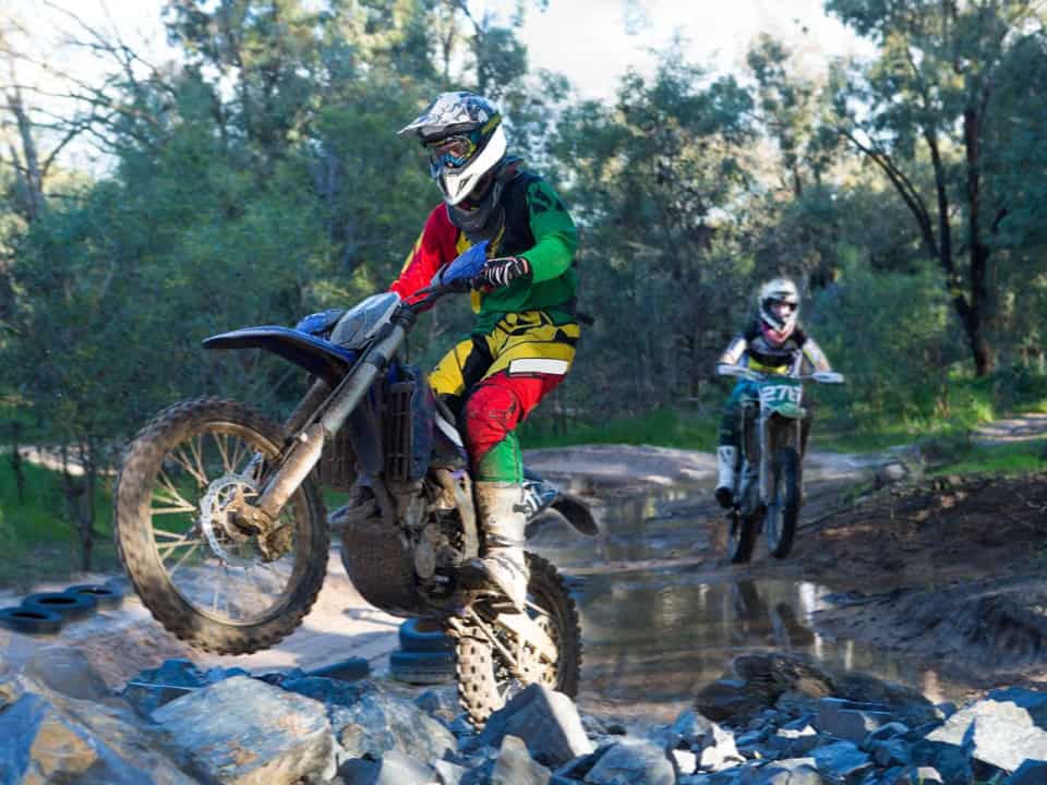 Two motocross riders navigate rocky terrain and water. The front rider is airborne, wearing green, yellow, and red gear. The second rider follows, wearing blue and white gear. Trees surround them.