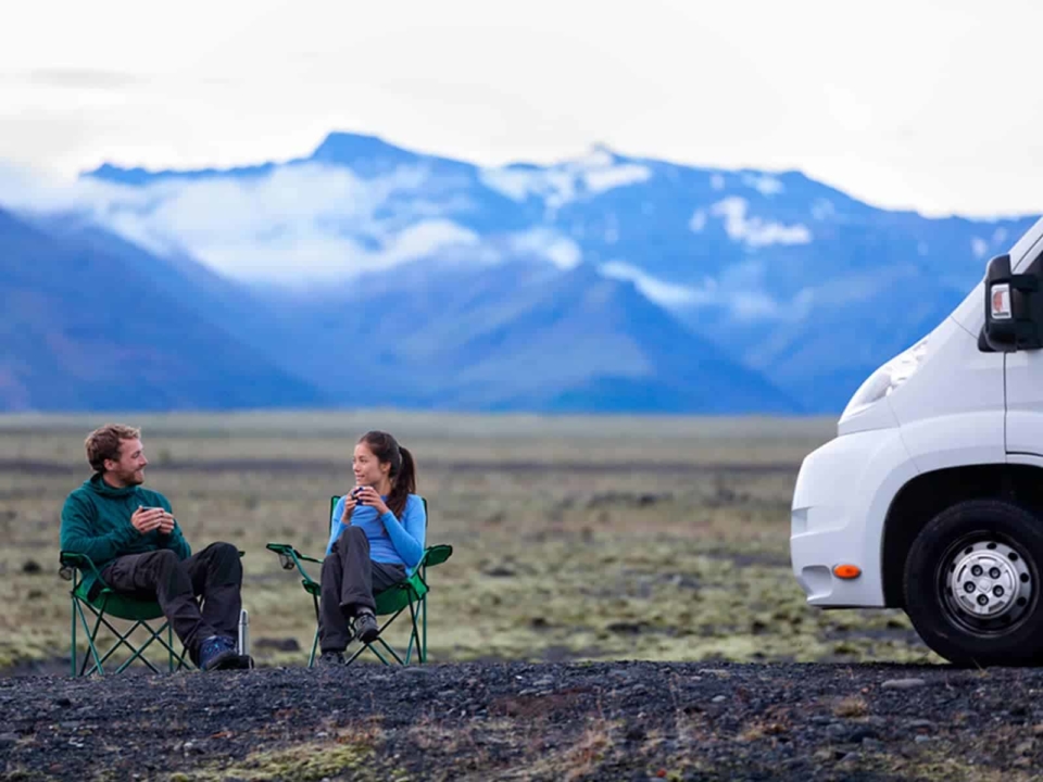 Couple drinking coffee next to their RV with snowy mountains in the background