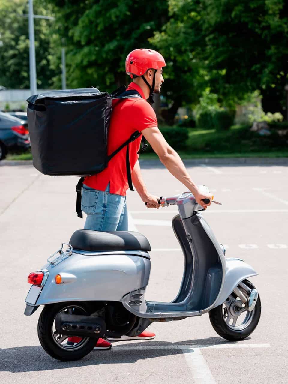 Person with a red helmet and shirt rides a scooter while carrying a large black delivery backpack.