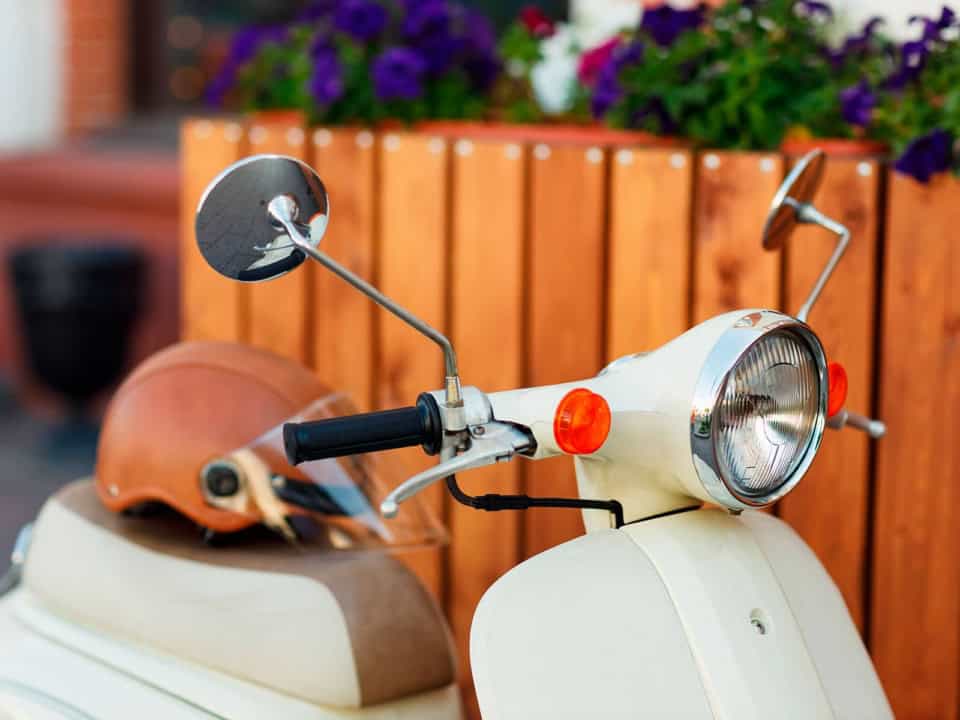 Close-up of a cream-colored scooter with a brown seat and helmet, featuring side mirrors and a headlight. Purple flowers in a wooden planter are in the background.