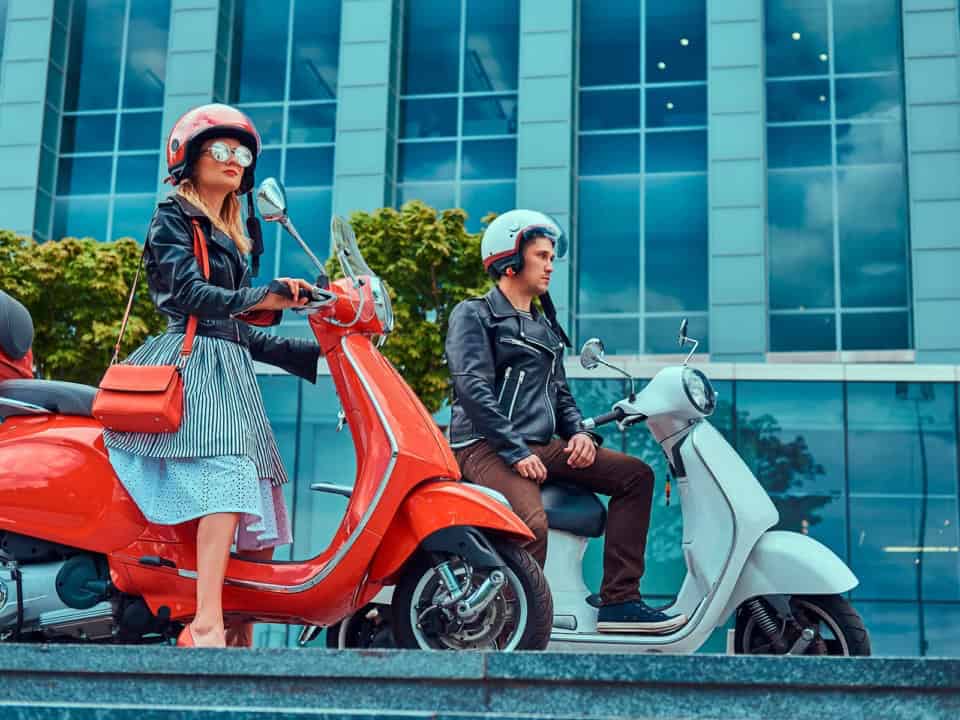 Two people wearing helmets sit on scooters, one red and one white, in front of a modern glass building.