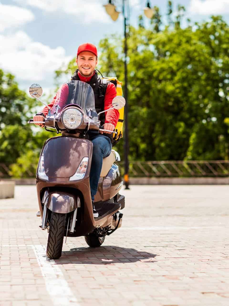 Person on a scooter wearing a red cap and backpack, riding on a paved path with green trees in the background.