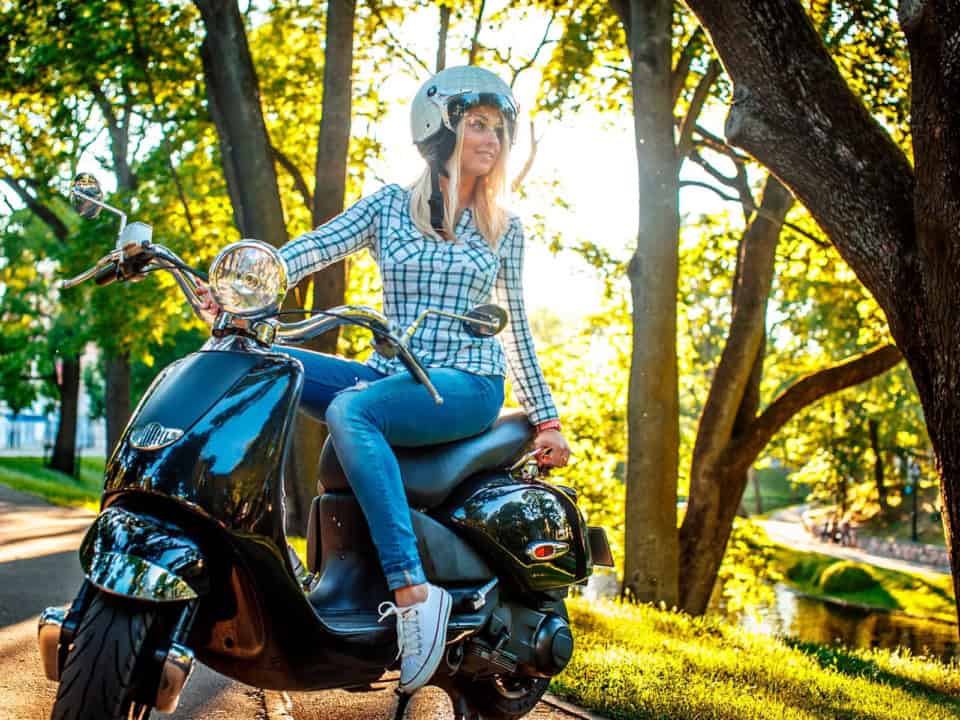 A woman wearing a helmet sits on a black scooter, surrounded by trees, with sunlight filtering through the leaves. She is dressed in a checkered shirt and jeans.