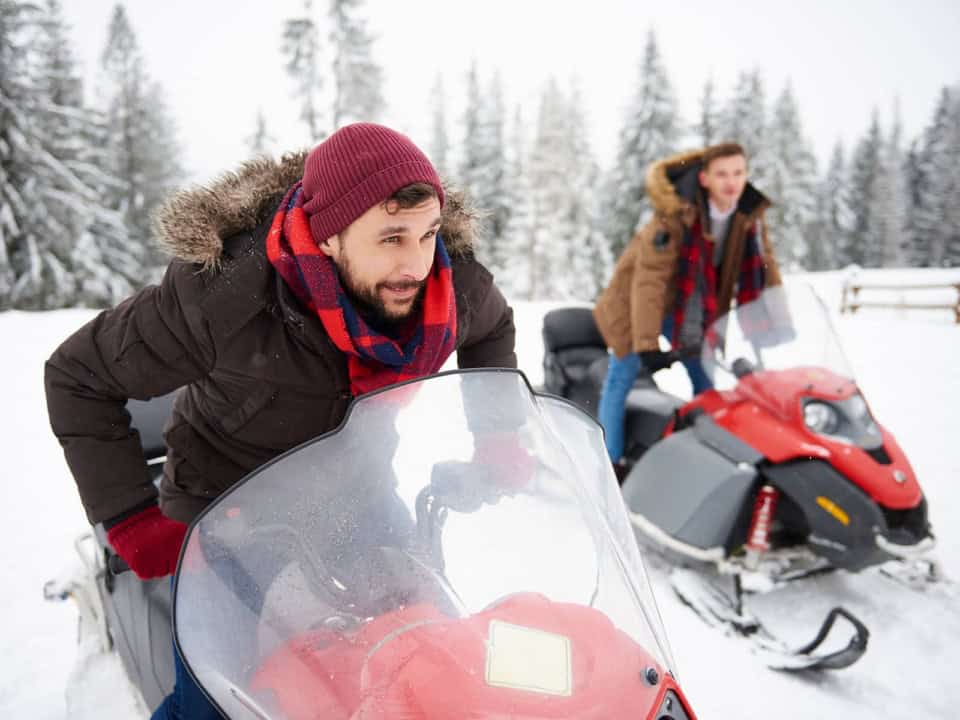 Two people in winter clothing prepare to ride red snowmobiles in a snowy forest setting.