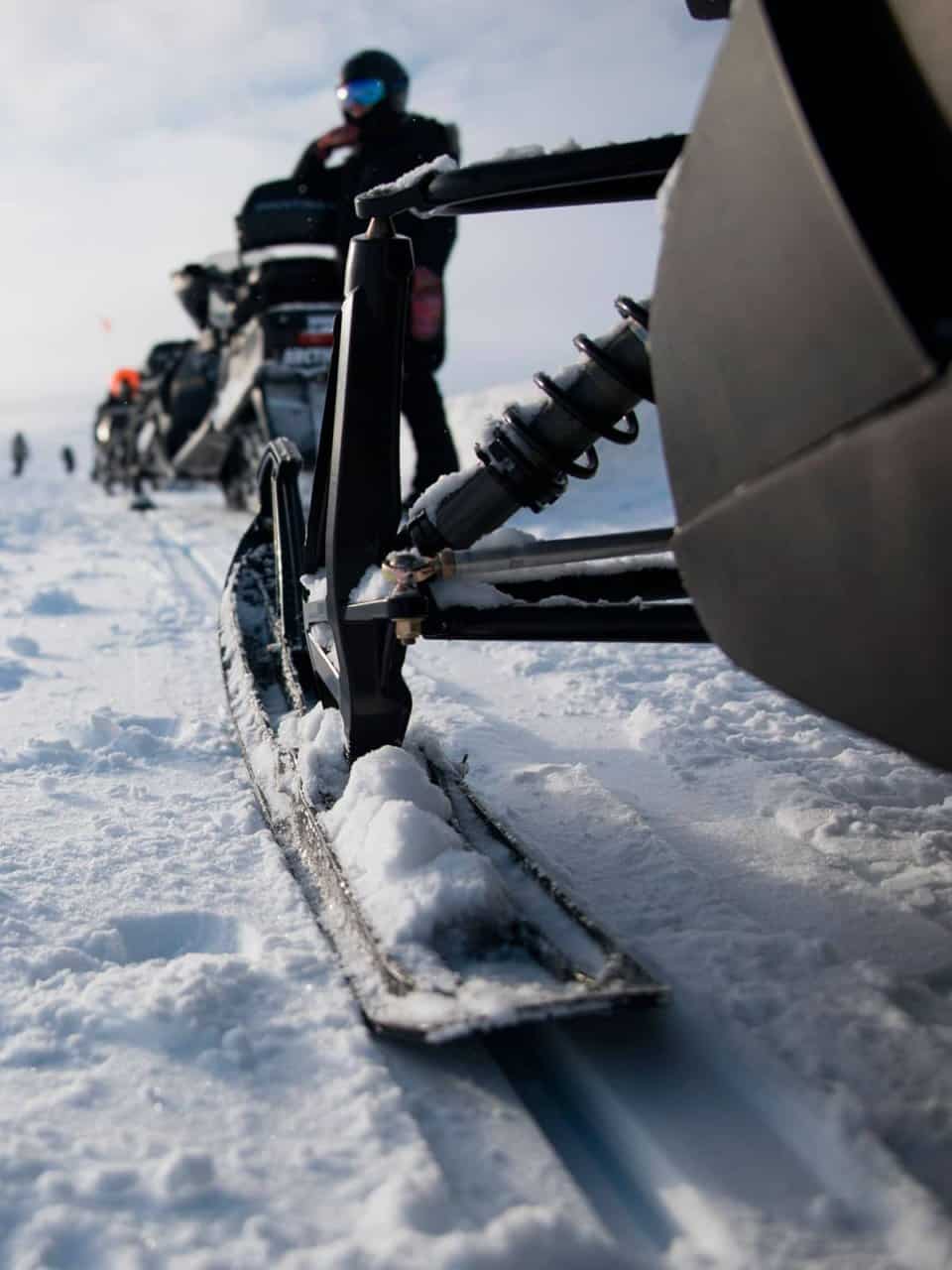 Close-up of a snowmobile ski on a snowy landscape with a blurred figure in the background wearing winter gear.