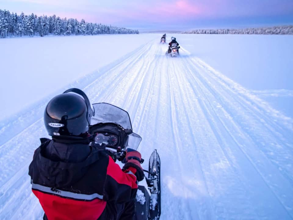 People riding snowmobiles on a snowy trail under a pinkish sky.