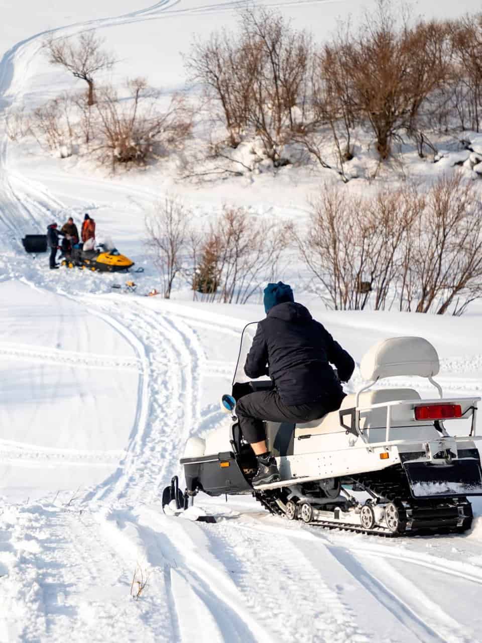 A person rides a snowmobile up a snowy hill. In the background, a group of people with another snowmobile and sled can be seen.