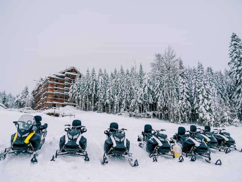 Line of snowmobiles parked in snowy landscape with a wooden building and snow-covered trees in the background.