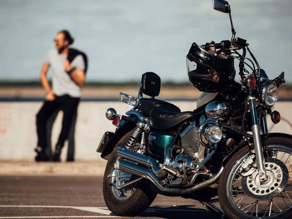 A parked motorcycle on a road in the foreground with a blurred background showing a person leaning on a railing.