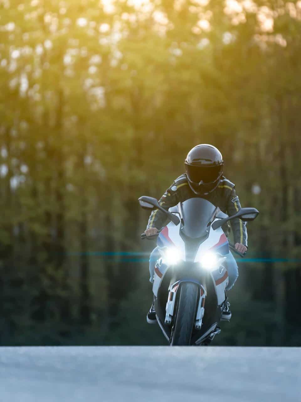 A person riding a motorcycle on a road with bright headlights, surrounded by a forest under a hazy sky.