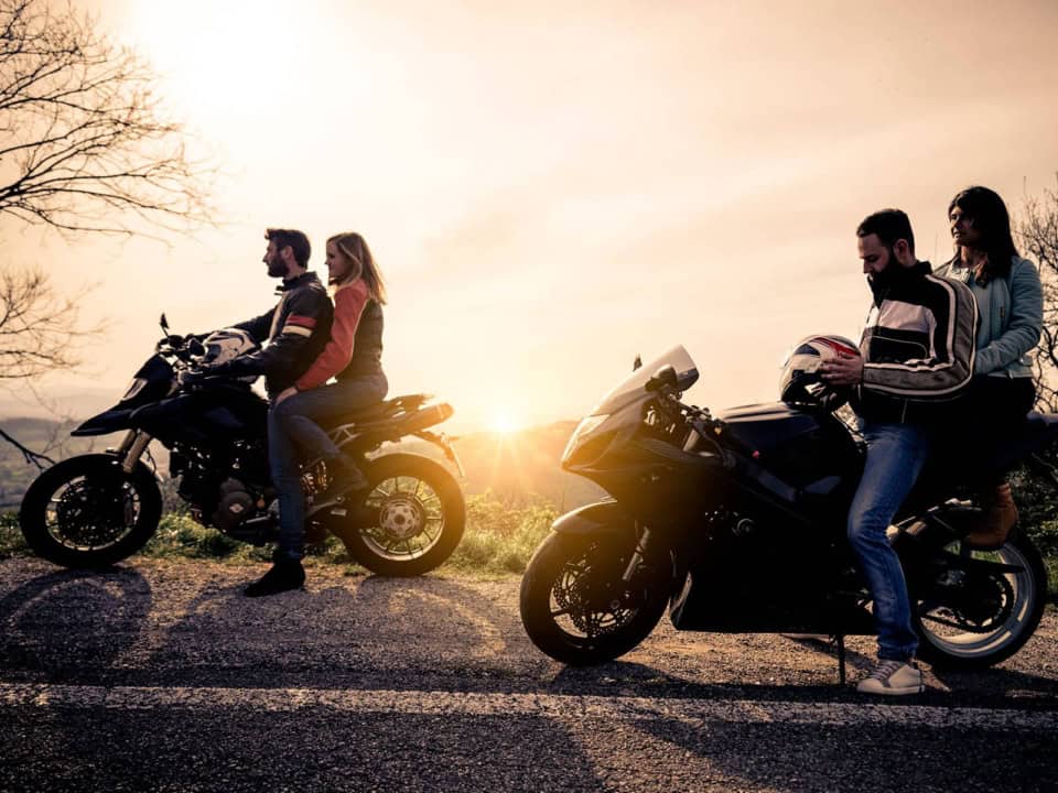 Two couples on motorcycles are parked by the roadside, with a sunset in the background.