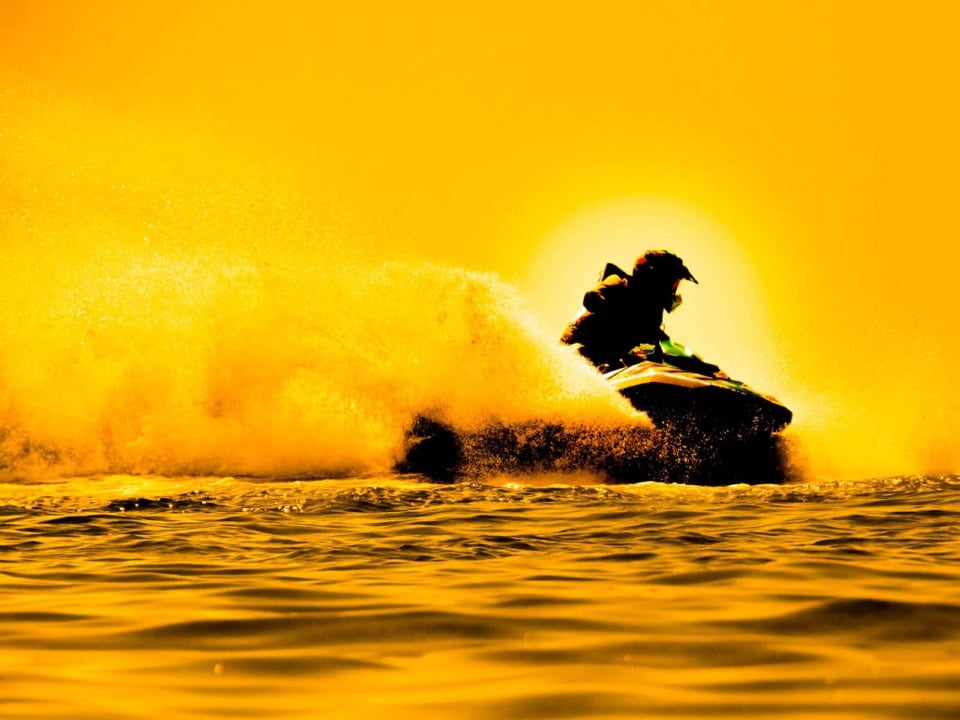 A person rides a jet ski on the water, creating a large splash, against a vibrant yellow sunset background.