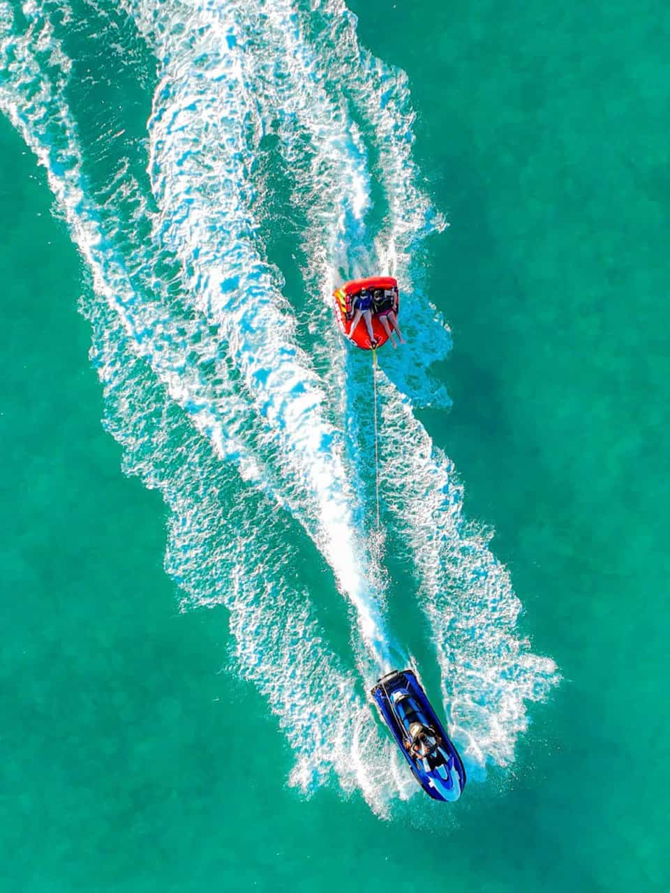 Aerial view of a jet ski towing a red inflatable tube with passengers over clear turquoise water, creating white wake trails.