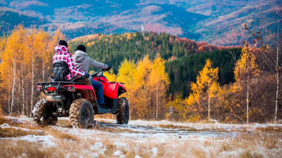 Two people on an ATV ride through a scenic autumn landscape with colorful trees and mountains in the background.