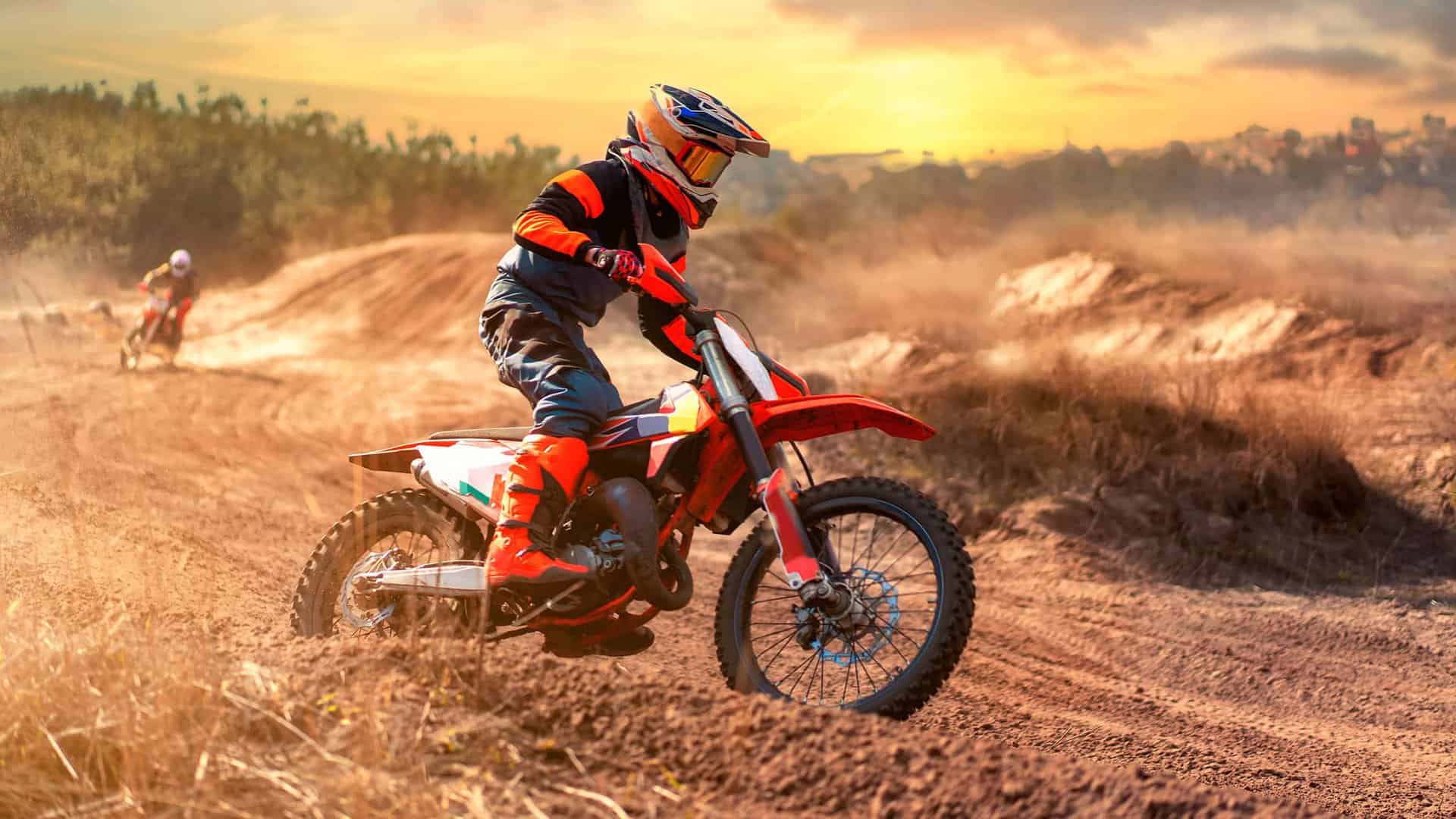 A motocross rider in red gear navigates a dirt track at sunset. Another rider is visible in the background. Dust clouds surround them.