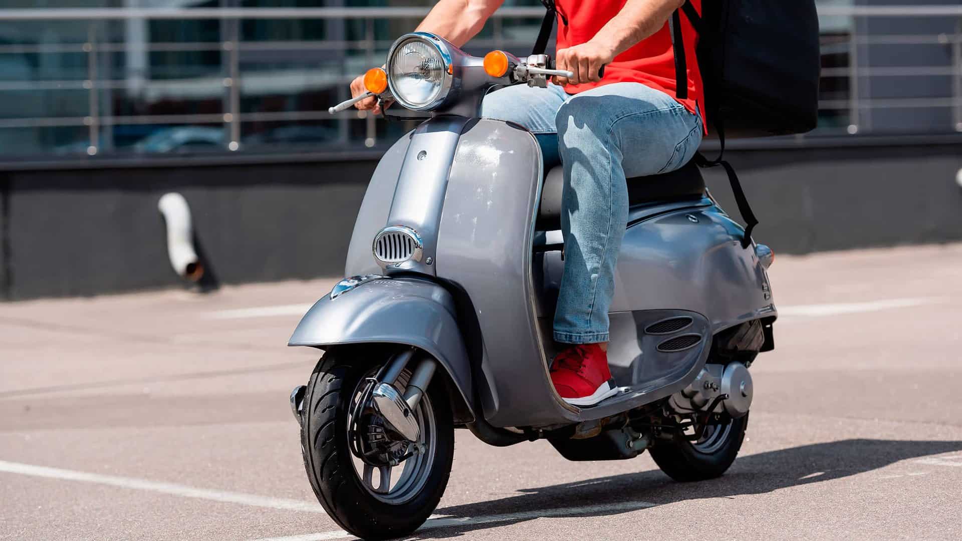 Person in casual attire riding a gray scooter in an outdoor area.