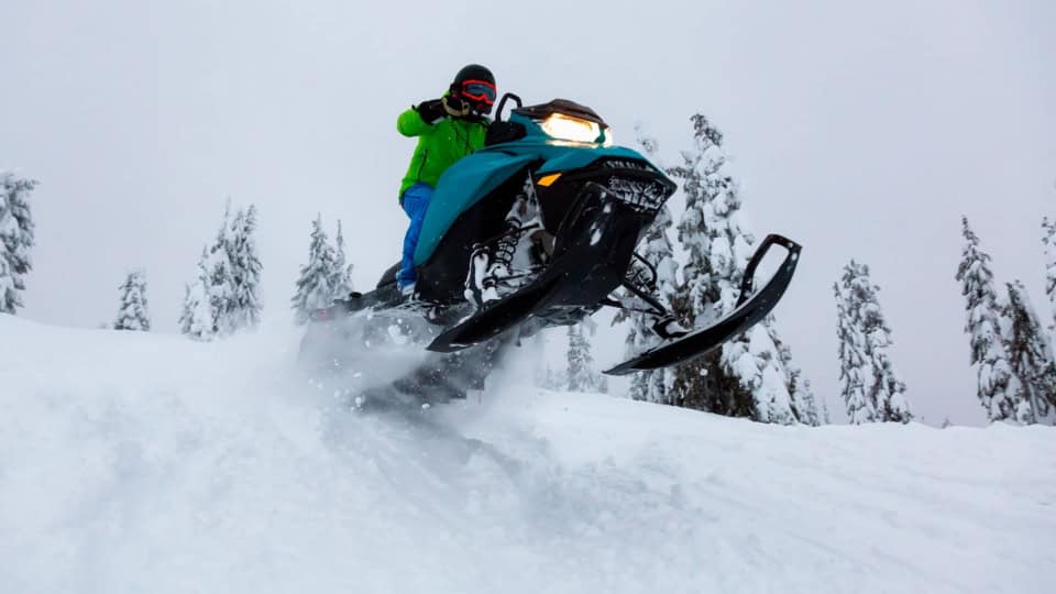 Person in a green jacket riding a snowmobile through snow-covered terrain, surrounded by trees.