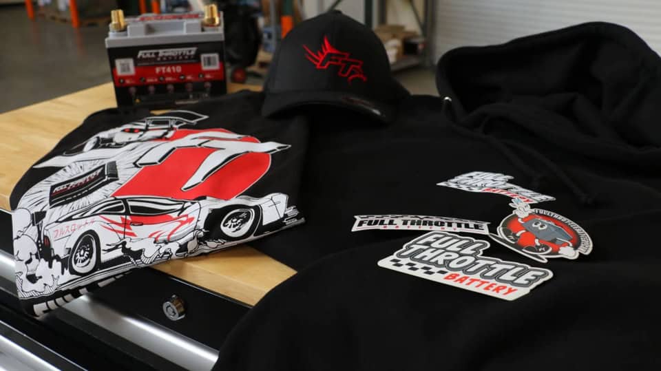 Various "Full Throttle" merchandise displayed on a table, including a battery, t-shirts, a cap, a hoodie, and stickers featuring racing car designs.